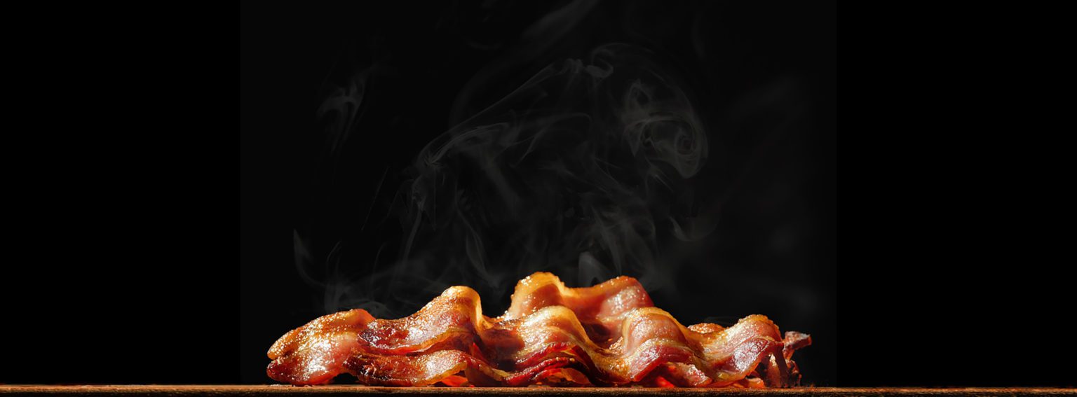 Crisp rashers of bacon with smoke rising on a dark background